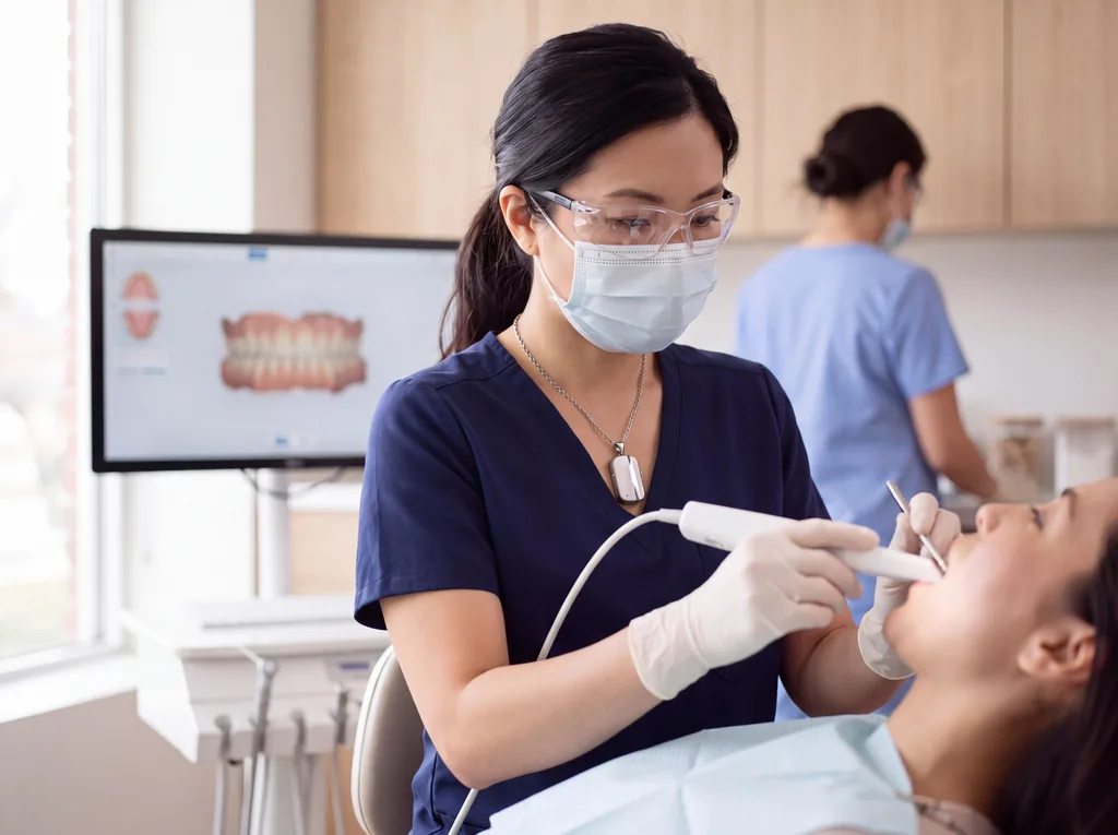 Illustration: a dentist wearing the Vault necklace takes an intraoral scan in a daylit operatory, the digital model on the screen behind her.